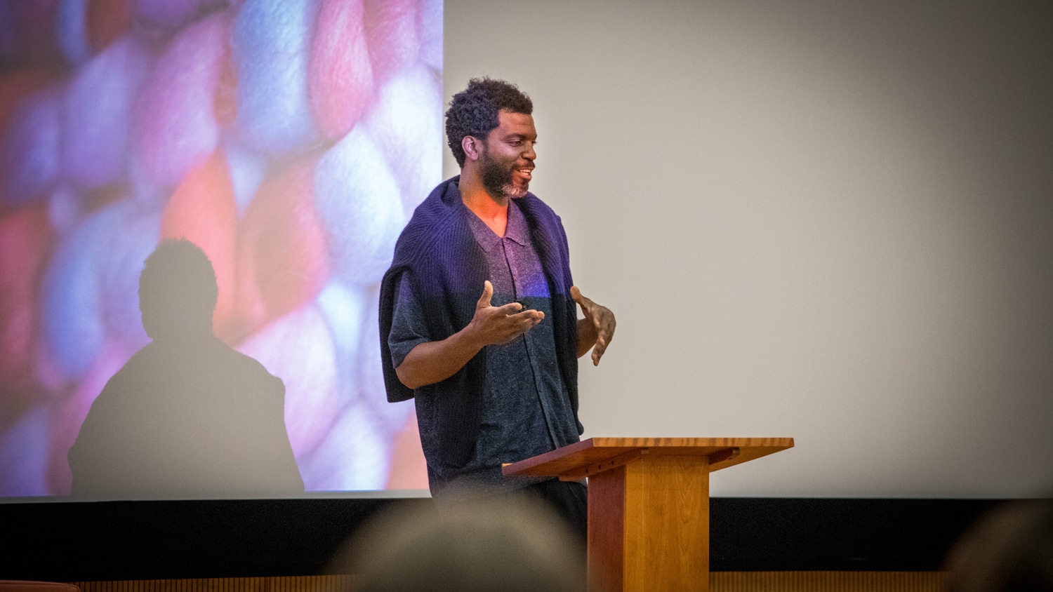 Stephen Satterfield stands at a wooden podium, gesturing while speaking. Behind him is a large, colorful abstract image projected on a screen. The audience is partially visible in the foreground.