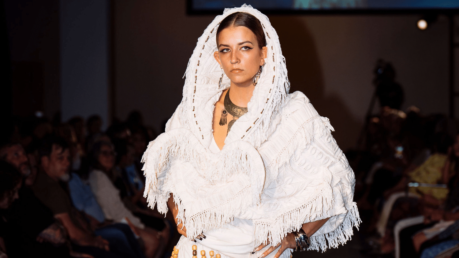 A model walks on a runway wearing a textured white outfit with fringe details and a hood. The garment features layered patterns and the model wears a statement necklace. The audience is visible in the dimly lit background.