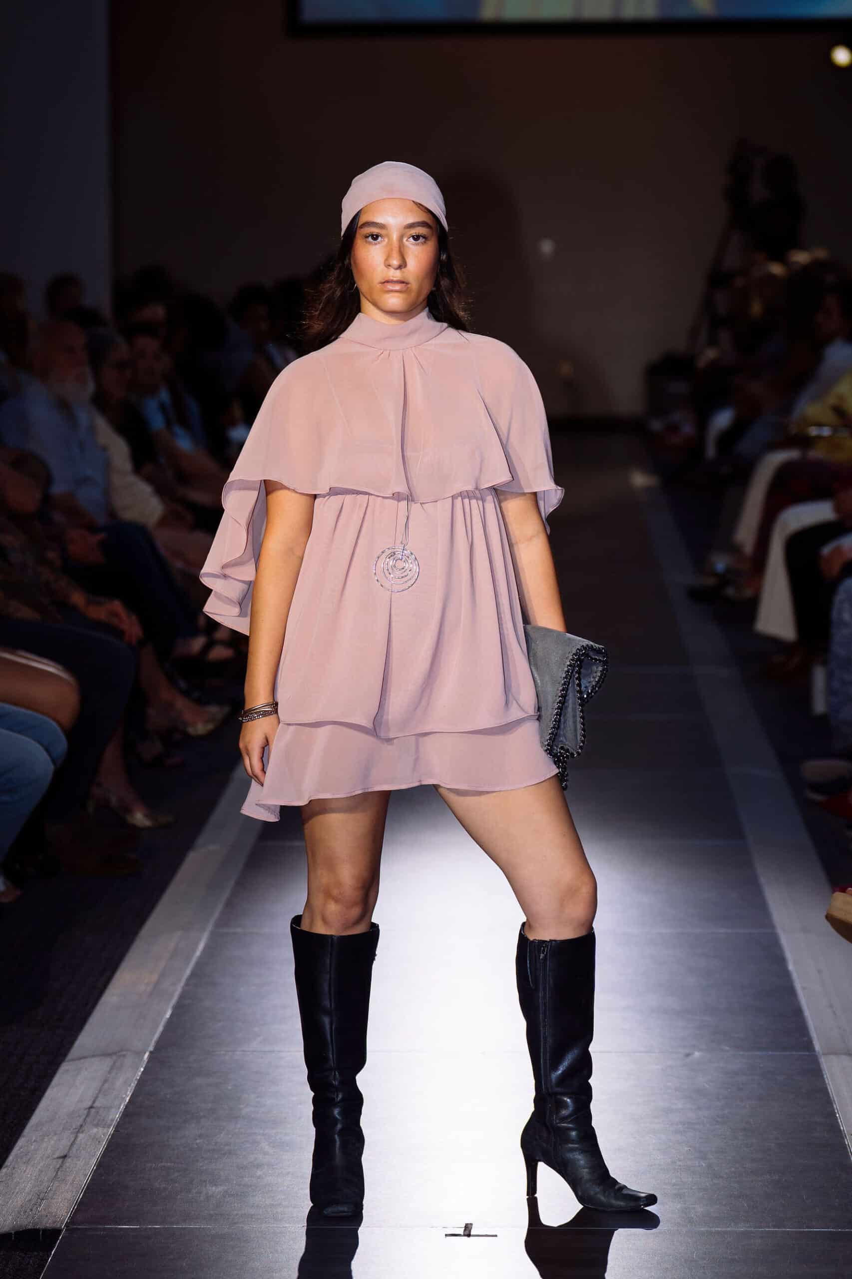 A model walks down a runway wearing a light pink, flowy dress with layered sleeves, a matching headband, black knee-high boots, and holding a black clutch. The audience sits along both sides of the runway.