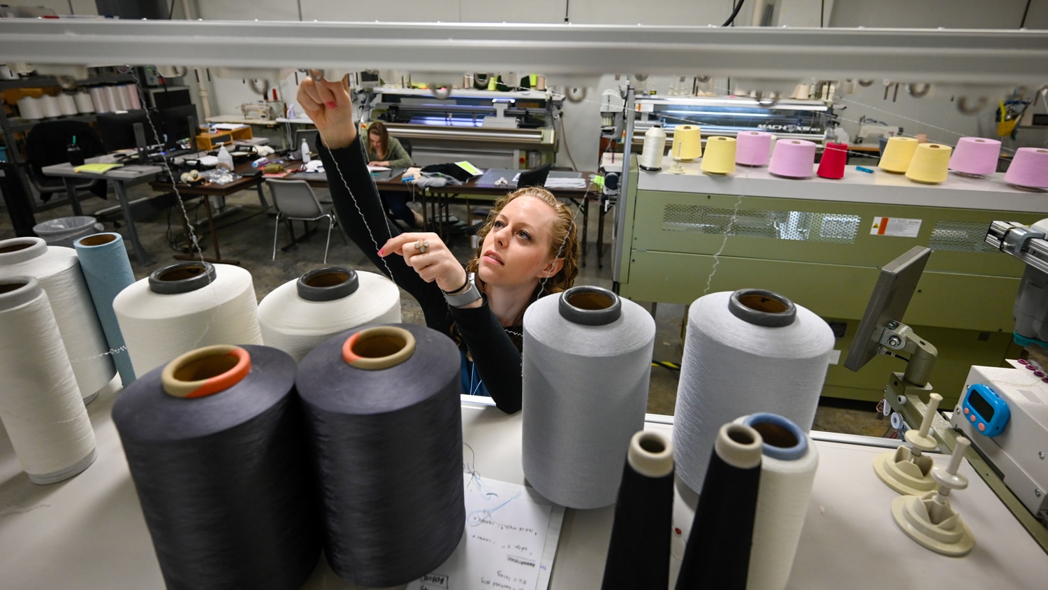 Zoe Hezrony works with thread spools on a machine in the Knitting Lab. She reaches up to adjust a thread with various large spools of different colors in the foreground and other knitting equipment in the background. Shayleigh Larsen is visible working at a table in the background.
