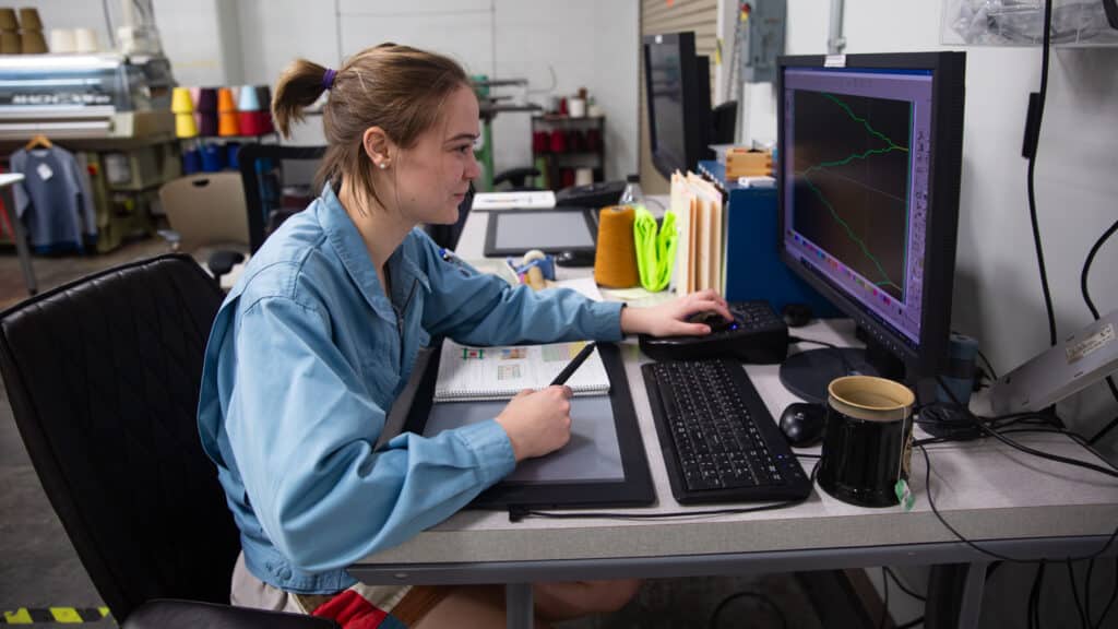 Shayleigh Larsen works at a desk with a drawing tablet, keyboard and computer monitor displaying colorful lines as part of a CAD file. She's in the Knitting Lab at NC State.