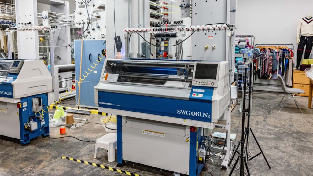A blue and white industrial knitting machine stands in the NC State Knitting Lab surrounded by other equipment and shelves, with racks of colorful clothing and yarn visible in the background.