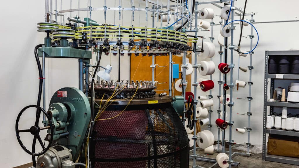 A circular industrial knitting machine with spools of white and red yarn on racks nearby, set up in the NC State Knitting Lab with shelves holding additional yarn supplies in the background.