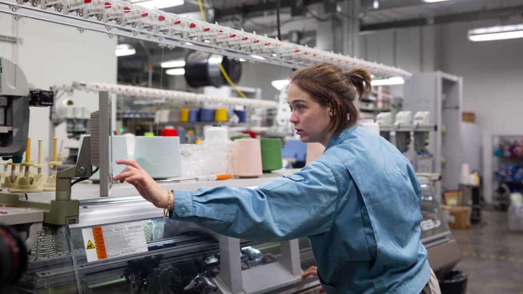 Shayleigh Larsen operates a machine in the Knitting Lab, reaching toward a control panel with spools of colorful yarn visible on the machine.