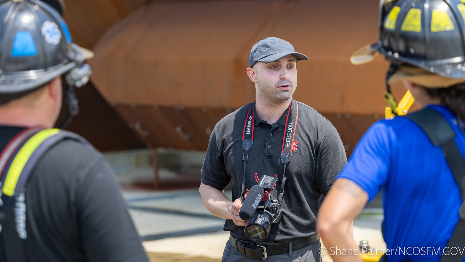 Arash Kasebi holds a camera and speaks to two people in firefighter helmets and gear outside near a large brown industrial structure.