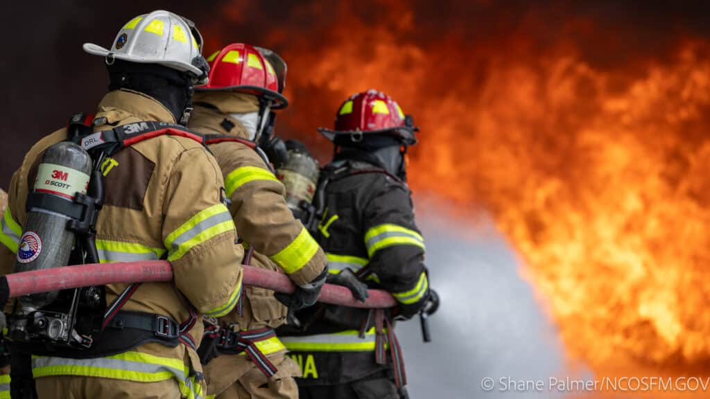 Three firefighters in full gear and helmets hold a fire hose, facing large, intense flames.