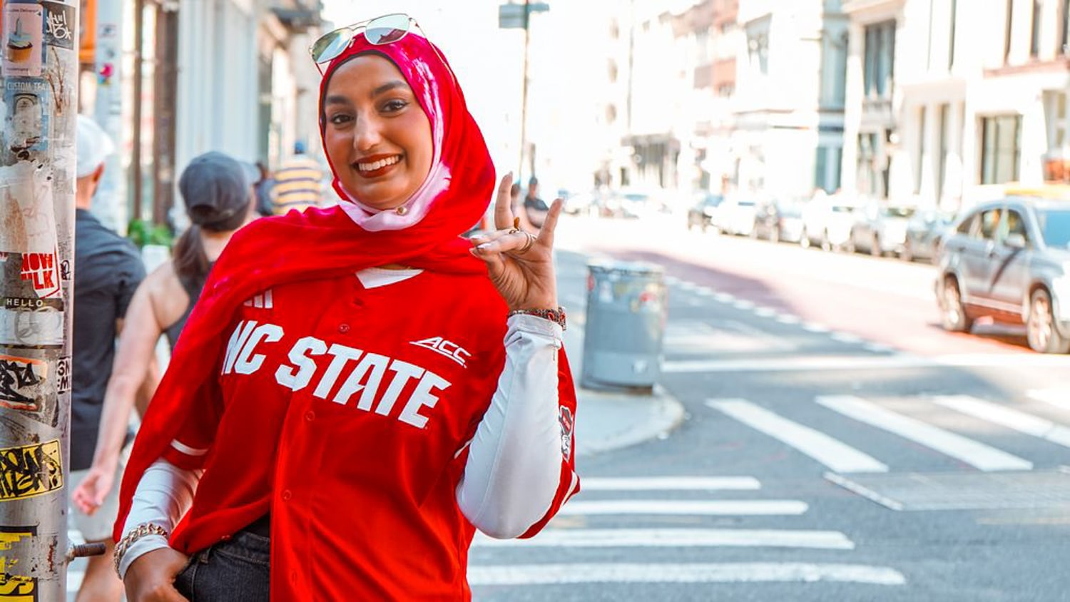 Humairaa Zafiruddin wears a red NC State jersey and a red and white hijab. She stands on a New York City sidewalk, holding up her hand in a wolf sign. She wears sunglasses on her head and several bracelets. The background shows cars and pedestrians.
