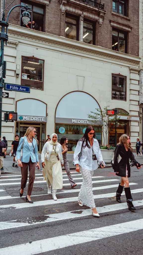 Humairaa Zafiruddin and four other people cross a street at a crosswalk in New York City, with a MEDRITE storefront and Fifth Ave street sign visible in the background.