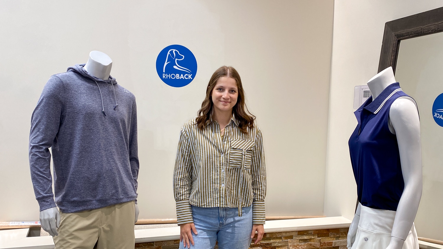 A woman stands between two mannequins dressed in sporty outfits inside the Rhoback office. A plush dog sits on the floor nearby, and a Rhoback logo is visible on the wall behind them.
