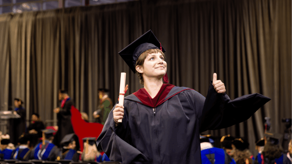 Wearing a cap and gown, Cecilia Monge holds a diploma and gives a thumbs up, smiling proudly at a commencement ceremony. Other graduates and faculty are visible in the background.