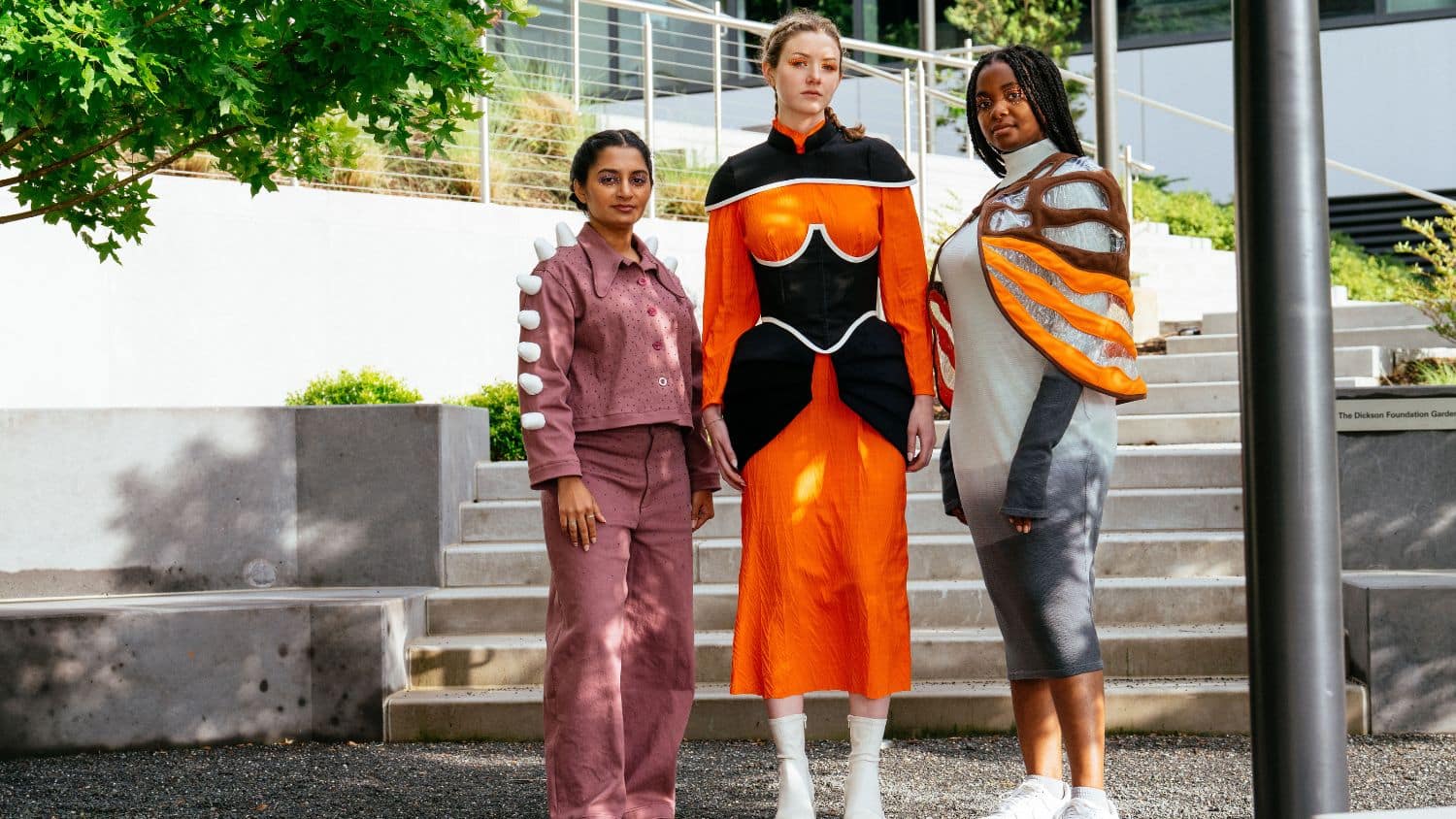 Three models stand outdoors on a gravel path, each wearing unique outfits in both earthy and bright tones, with architectural stairs and greenery in the background.