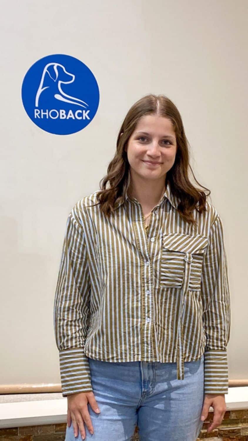 A woman stands in an office next to a Rhoback company logo. 