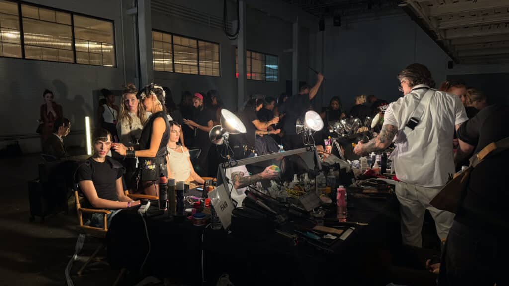 Behind-the-scenes view of a busy backstage area at a fashion show, with makeup artists working on models seated at tables covered in beauty supplies, all illuminated by bright vanity lights.