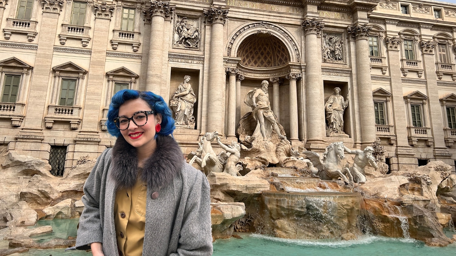 Athalia Windham, wearing a gray coat with a fur collar, stands smiling in front of the Trevi Fountain during her spring break trip to Rome, Italy.