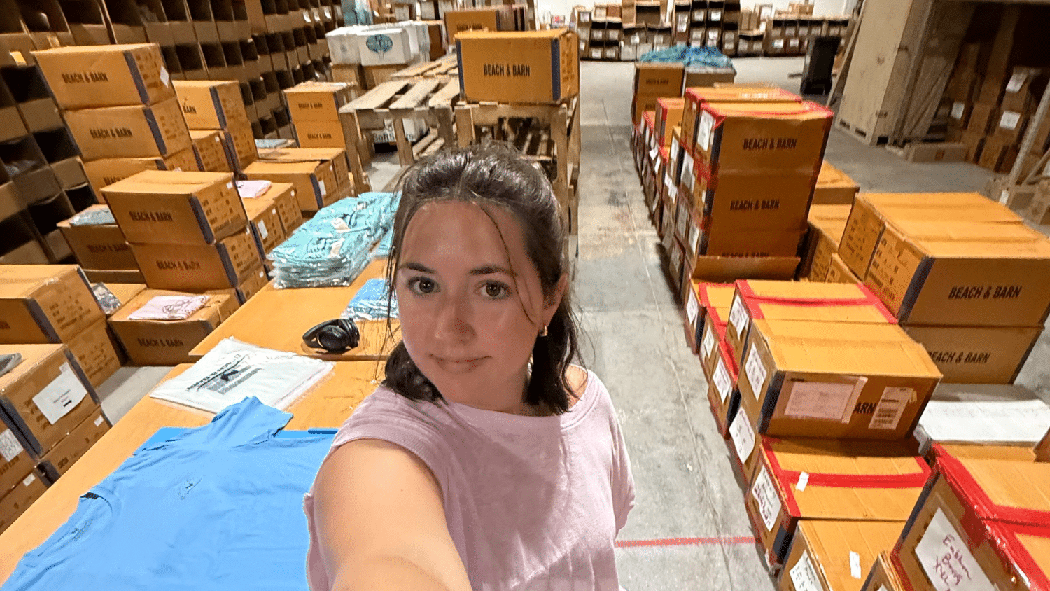 Georgia Allen takes a selfie in a warehouse filled with stacked boxes and folded shirts on tables, surrounded by shelves stocked with more boxes.