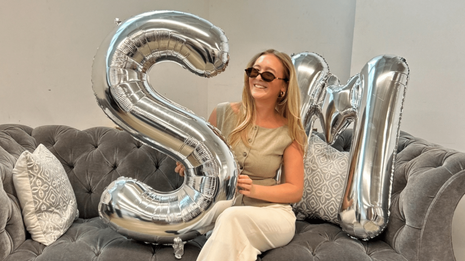 Giada Cavaliere wears sunglasses and a beige top while sitting on a gray couch, smiling and holding large silver letter balloons shaped like "S" and "M." Patterned pillows are on the couch beside her.