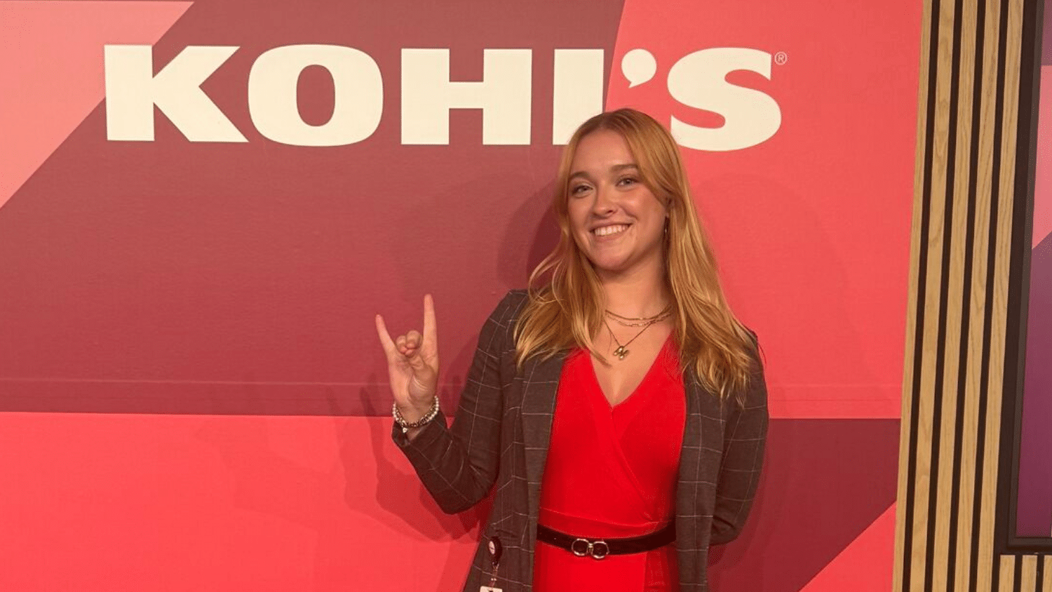 Nikki Fiske wears a red dress and blazer standing in front of a red backdrop with the "KOHLS" logo, holding up the Wolfpack hand gesture.