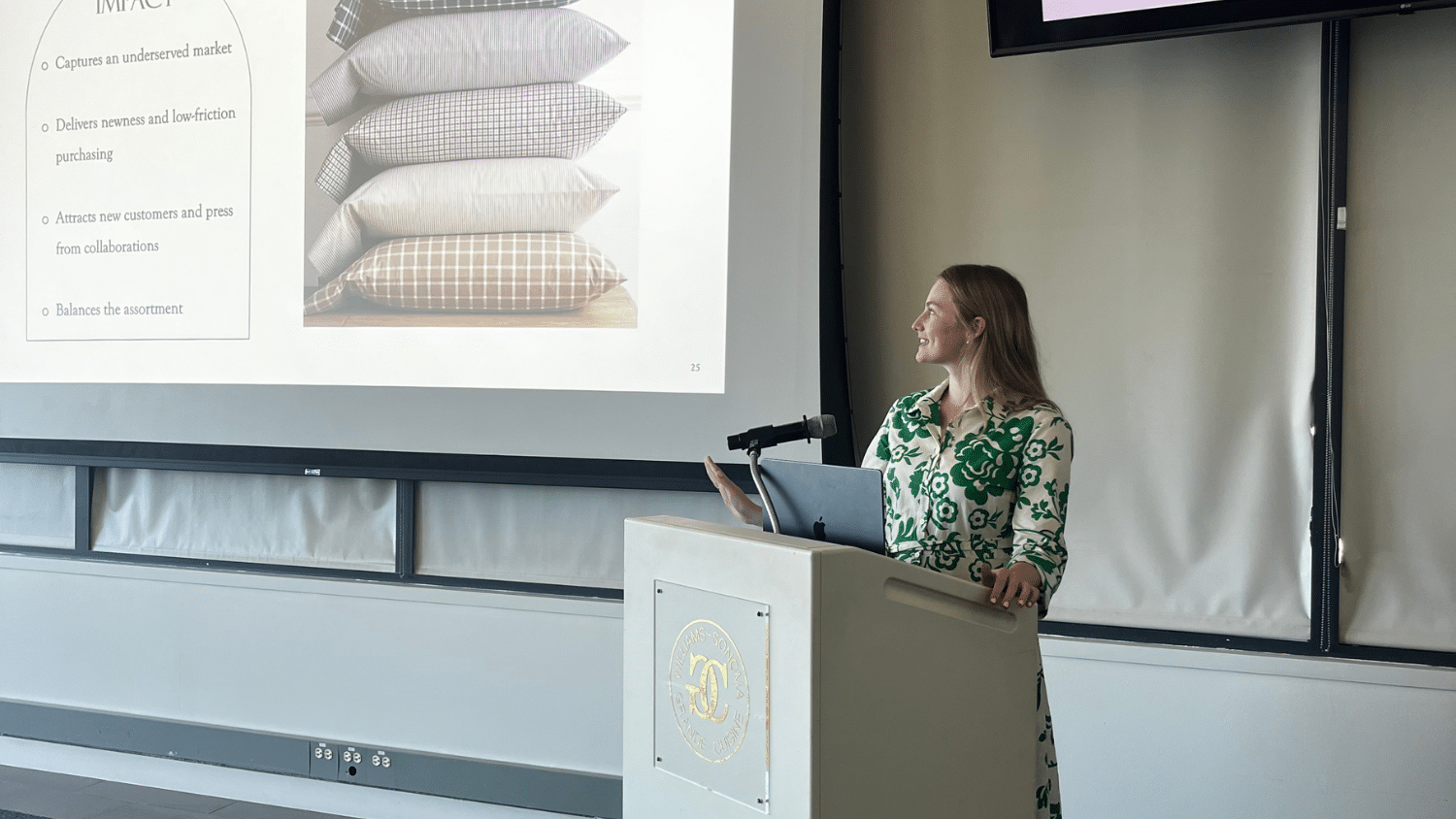 Luisa Warlick stands at a podium, presenting to an audience. She gestures toward a projected slide showing a stack of pillows and a bulleted list of points.