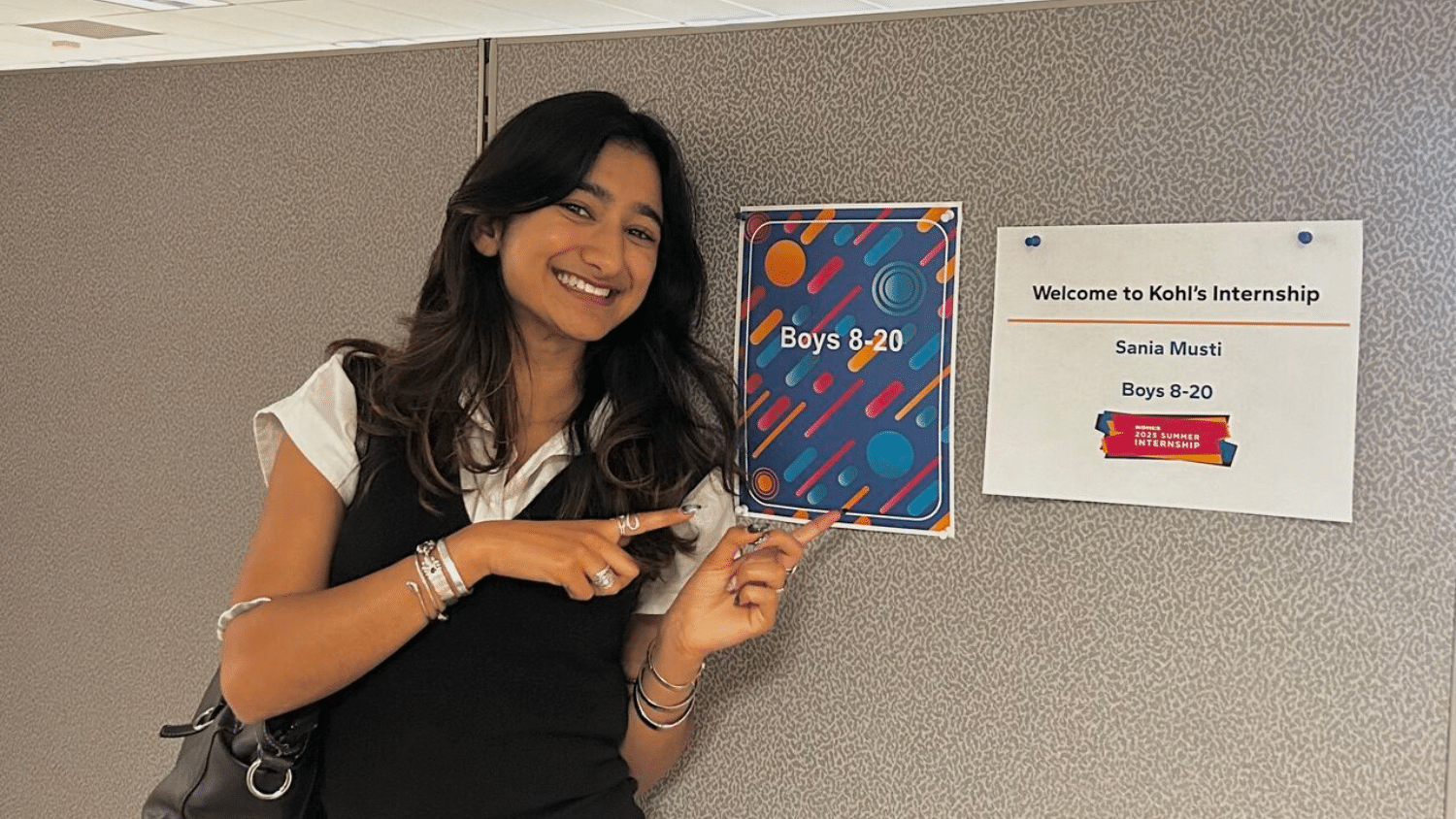 Sania Musti stands smiling indoors, pointing to a colorful sign that reads "Boys 8-20" on a wall. Next to it is a printed sheet that says "Welcome to Kohl’s Internship" with her name on it.
