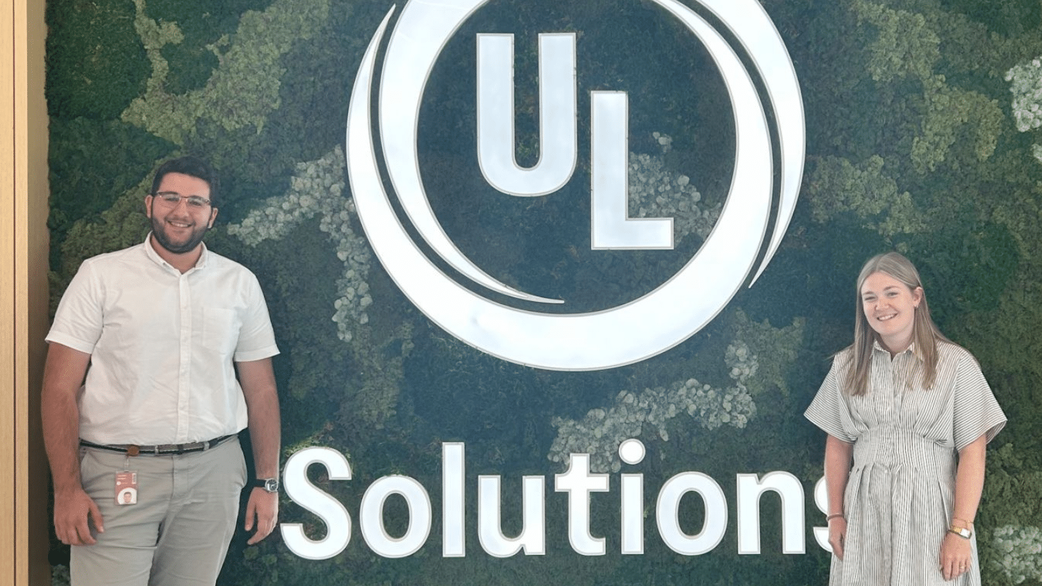 Youssef Kozman and fellow intern stand smiling in front of a large green wall featuring the "UL Solutions" logo in white. The person on the left wears a white shirt and dark pants; the person on the right wears a light-colored dress.