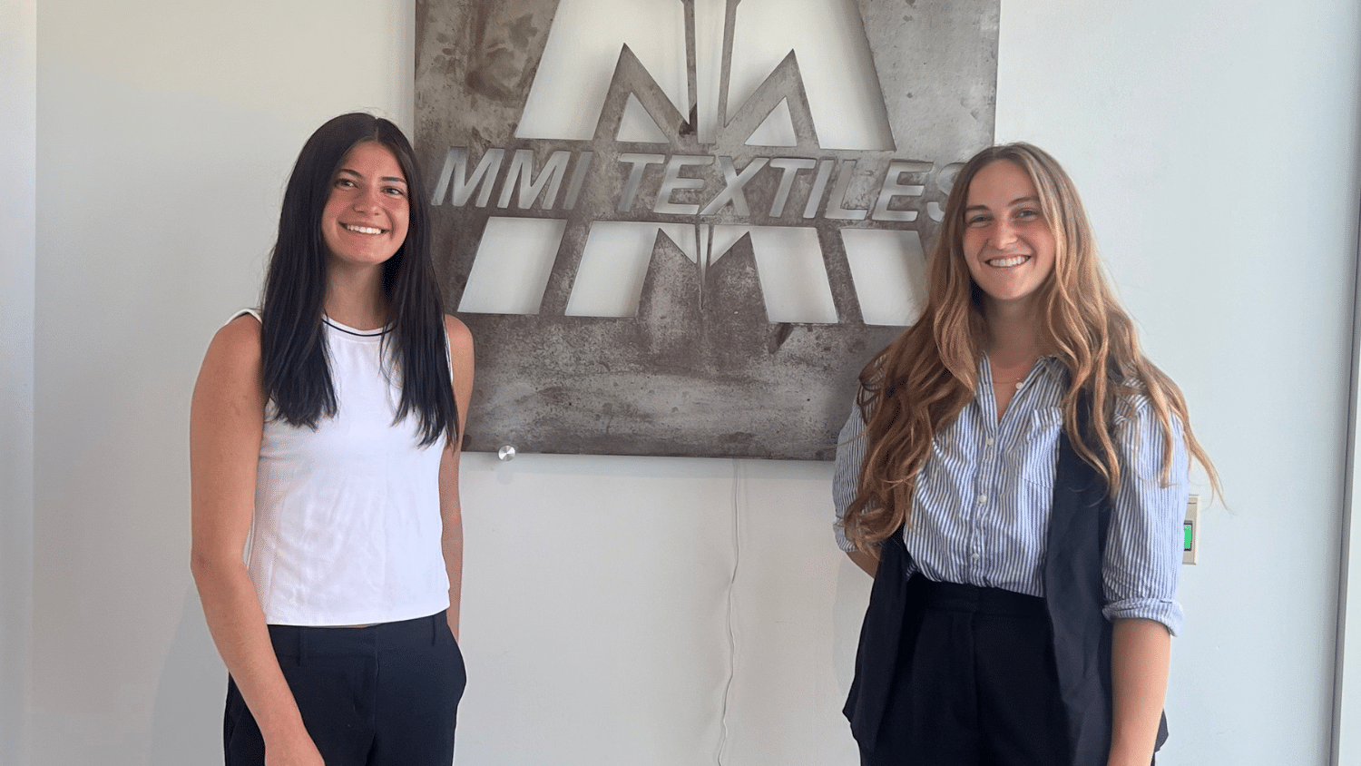 Mikayla Ruhland and fellow intern stand smiling in front of a metallic MMI Textiles sign on a white wall. Both are dressed in business casual attire.