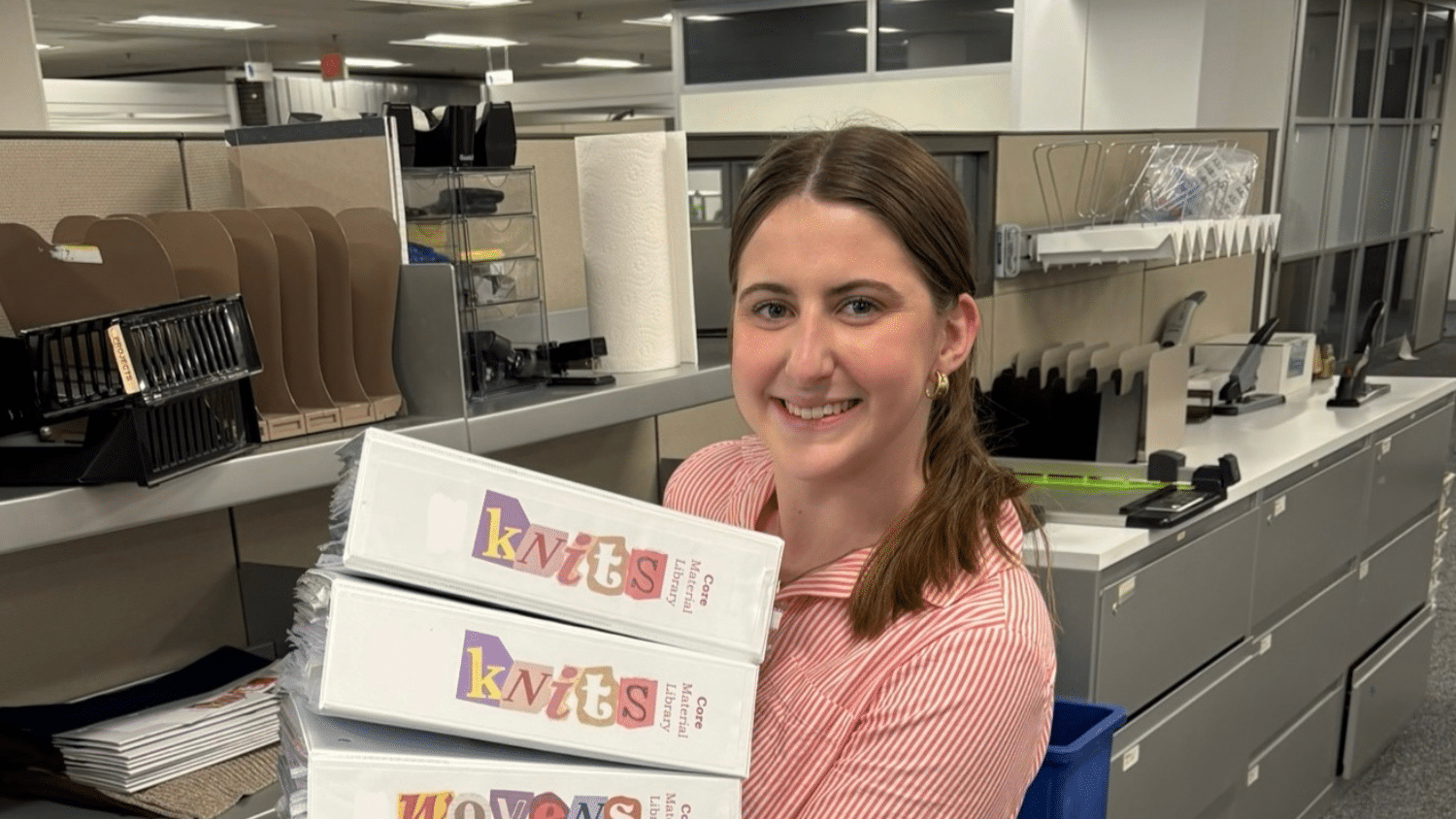 Annie Cameron smiles while holding three large binders labeled "Knits" in an office with cubicles, filing trays, and shelves in the background.