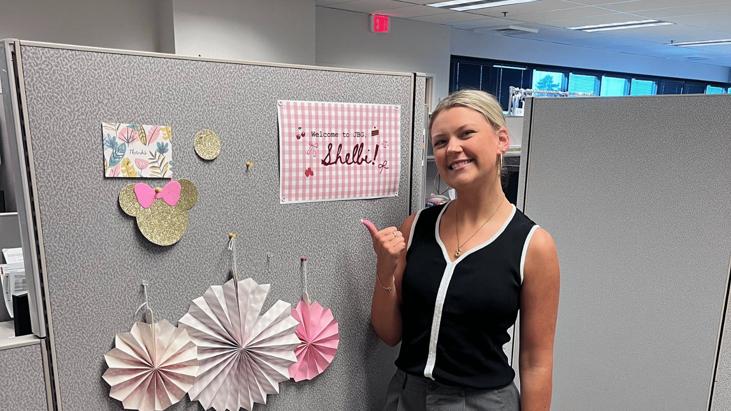 Shelbi Coffey stands smiling and pointing at a decorated office cubicle wall with paper fans, a mouse-shaped cutout, and a sign that says "Hello!"