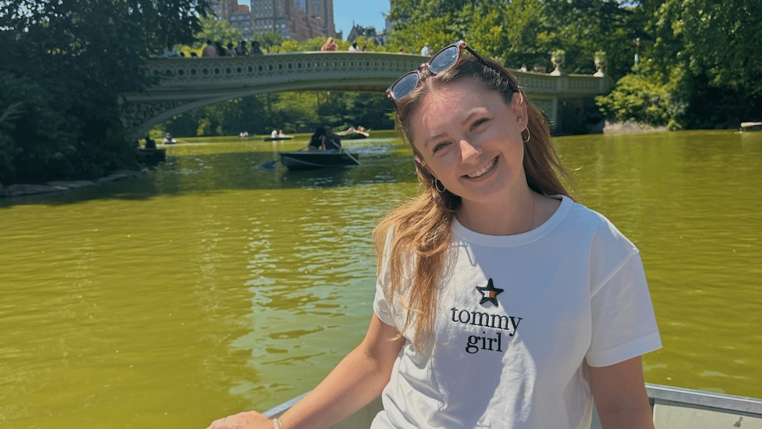 Norah Philbin wears a white "Tommy Girl" t-shirt while sitting in a rowboat on a greenish lake, with a bridge, trees, and people in the background on a sunny day.