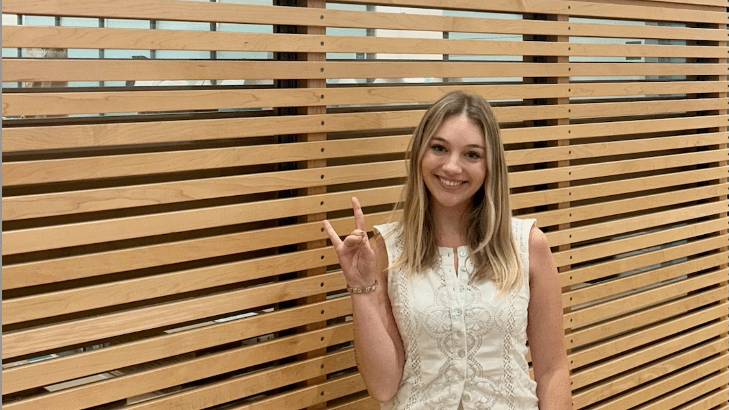 Bella Cybulski smiles at the camera while making the Wolf Pack hand gesture. She stands in front of a wall with horizontal wooden slats.