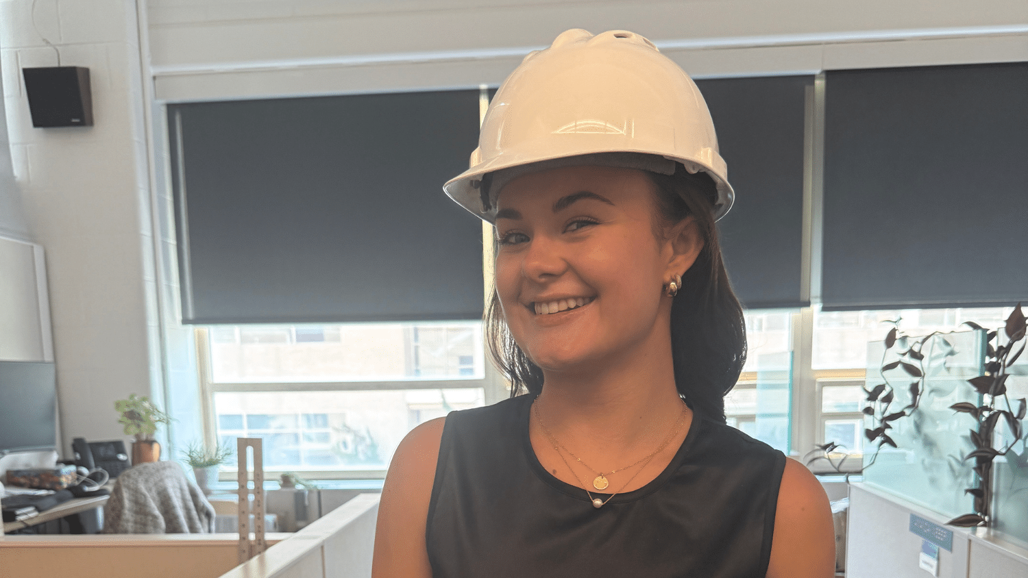Sophia Thompson wears a white hard hat and a sleeveless black top while smiling in a brightly lit office with large windows and plants in the background.