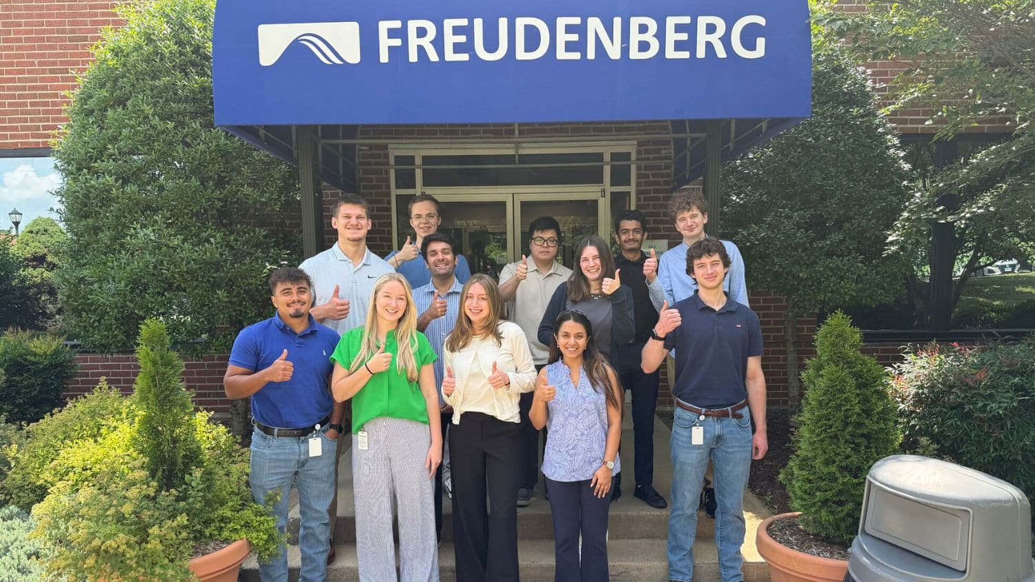 Mayuri Thakur and her fellow interns stand smiling and giving thumbs up in front of a building entrance with a blue awning labeled “FREUDENBERG.” Trees and bushes frame the entrance.