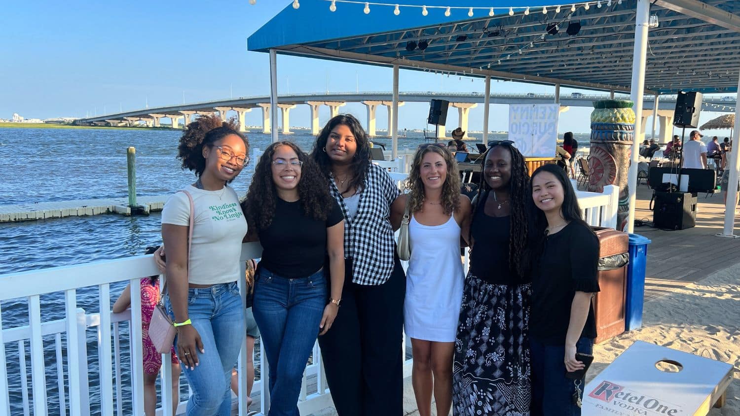 Mariella Neri and five others stand together smiling on a waterfront patio with a bridge and blue sky in the background. The group is dressed in casual summer attire and appears to be enjoying a sunny day by the water.