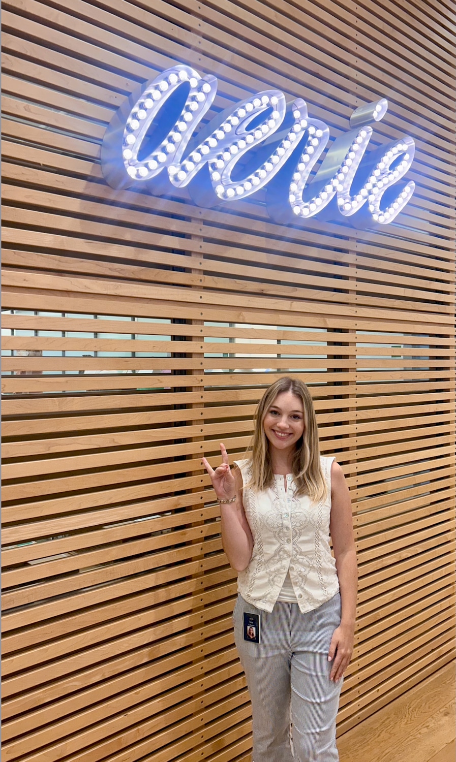 Bella Cybulski smiles at the camera while making the Wolf Pack hand gesture. She stands in front of a wall with horizontal wooden slats. There's also an Aerie sign made of lightbulbs.