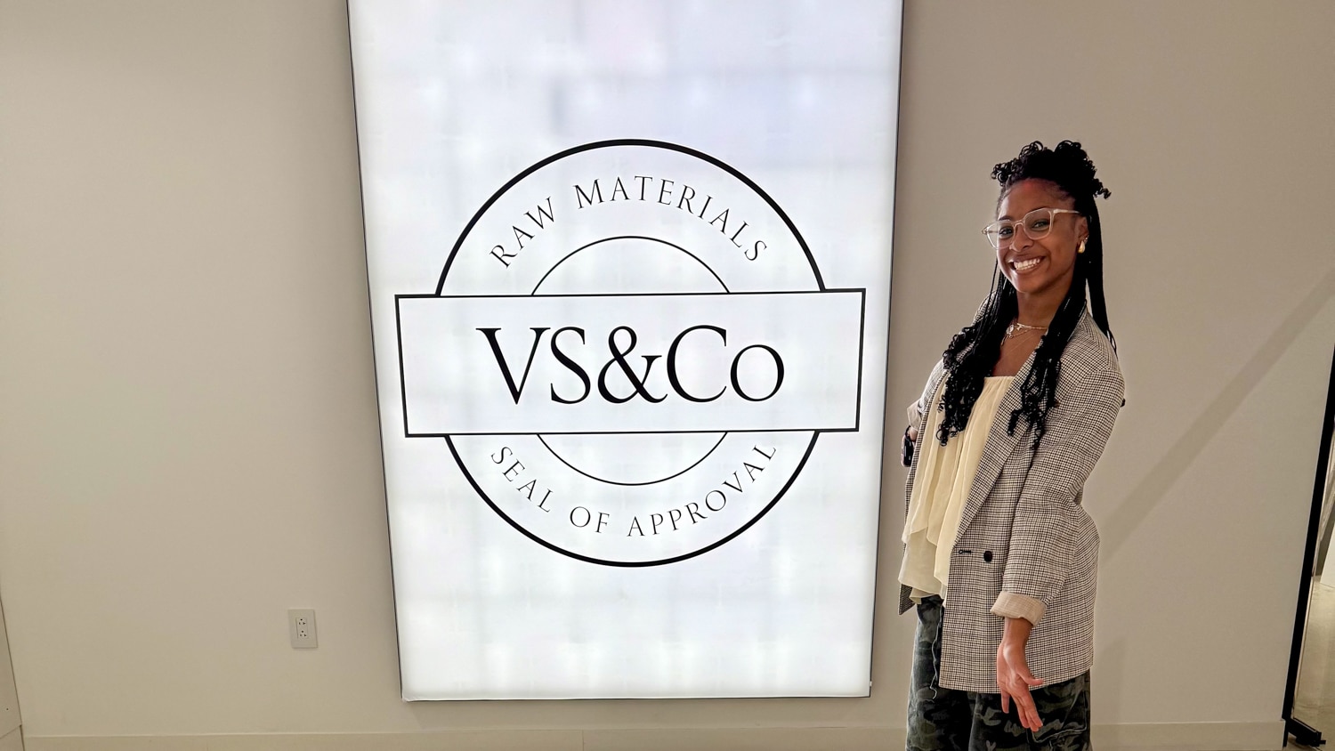Naomi Southerland poses in front of a large illuminated sign that reads "RAW MATERIALS SEAL OF APPROVAL VS&Co." The setting appears to be indoors with a modern, clean design.