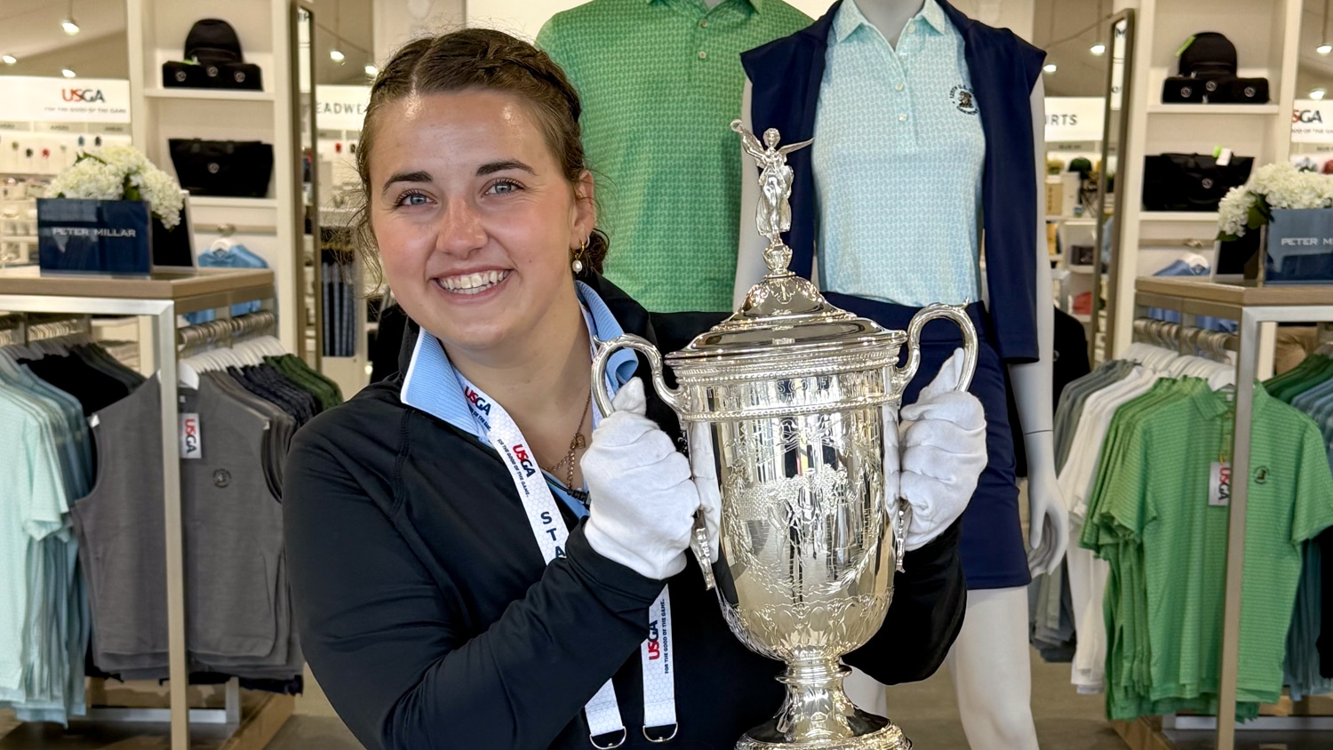 Rachel Whitaker holds a large silver trophy in front of two mannequins dressed in golf shirts at a merchandise store. She is wearing a black jacket and a lanyard around her neck.