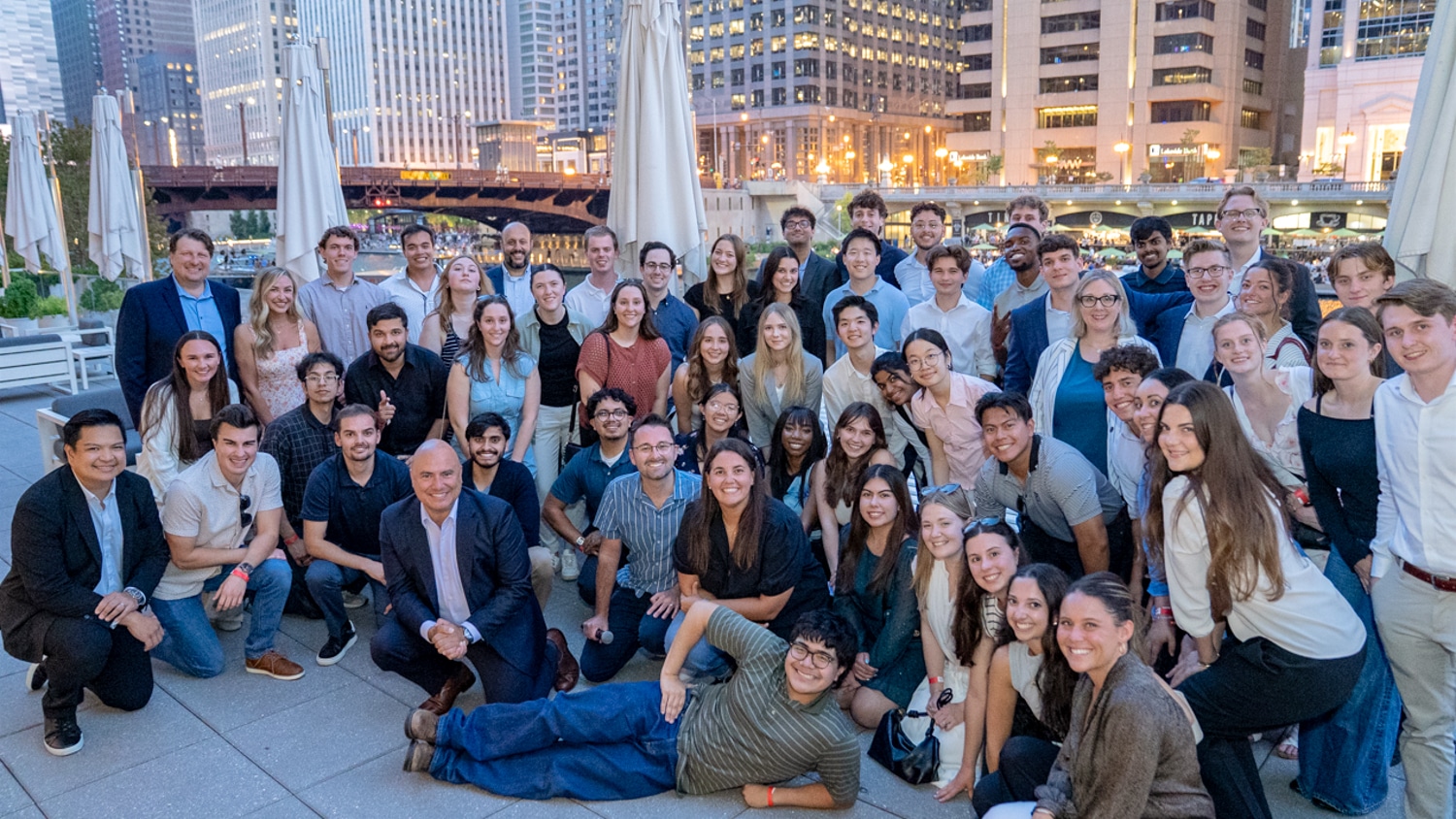 A large group of people pose and smile for a photo on an outdoor patio in a Chicago at dusk, with tall buildings and lights in the background. Some are standing, others are kneeling or sitting in the front.
