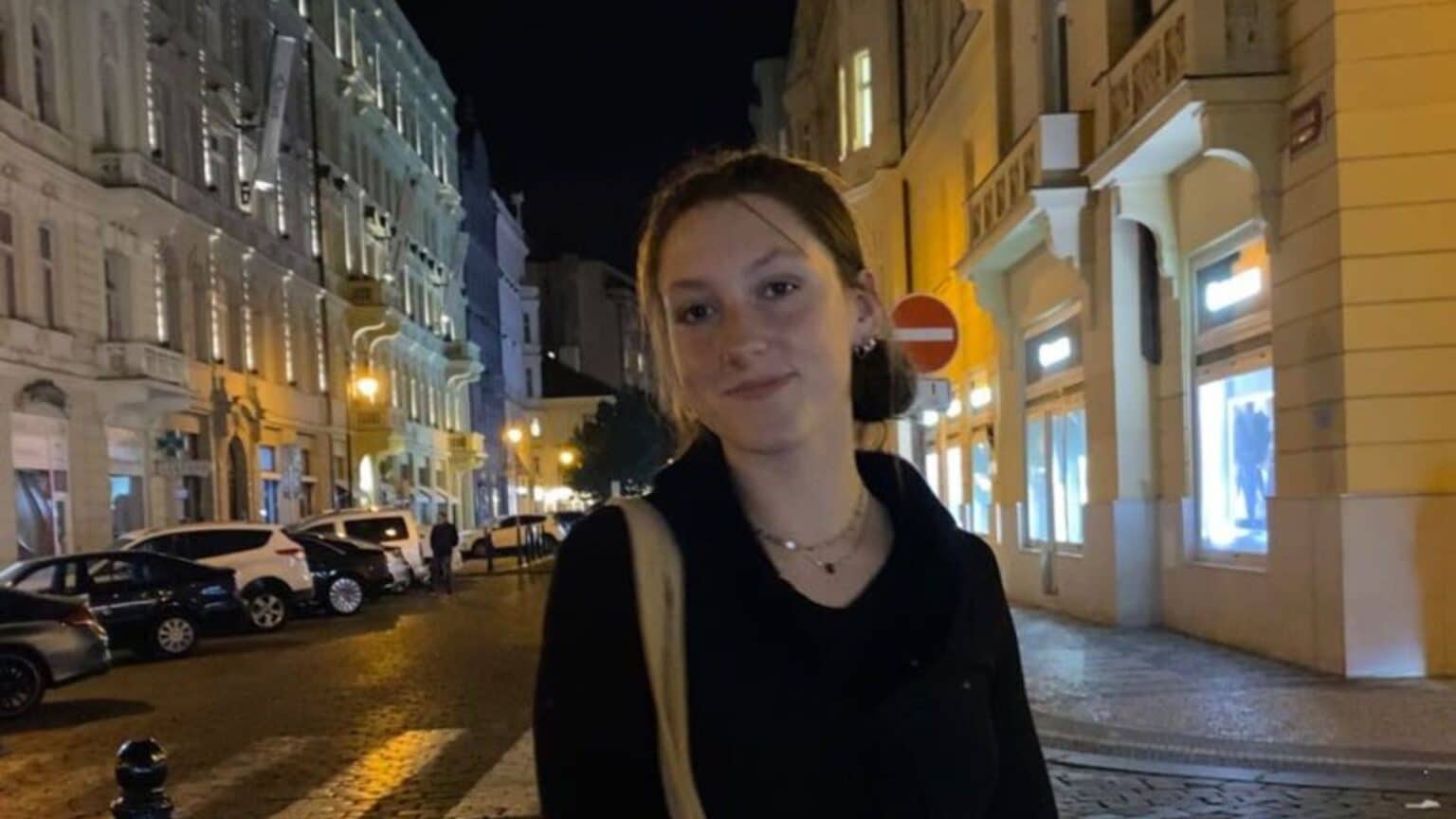 Rowan Mladsi stands at an intersection in Prague, Czech Republic at night. She is carrying a tote bag and smiling at the camera with lines of buildings and cars lit up behind her.