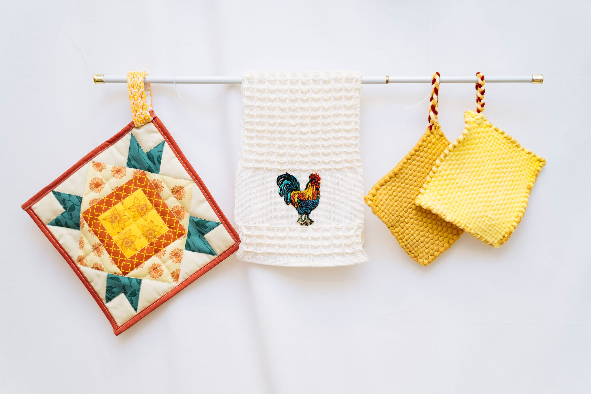 A colorful quilted potholder, a white towel with an embroidered rooster, and two yellow knitted potholders hang on a white rod against a plain white background.