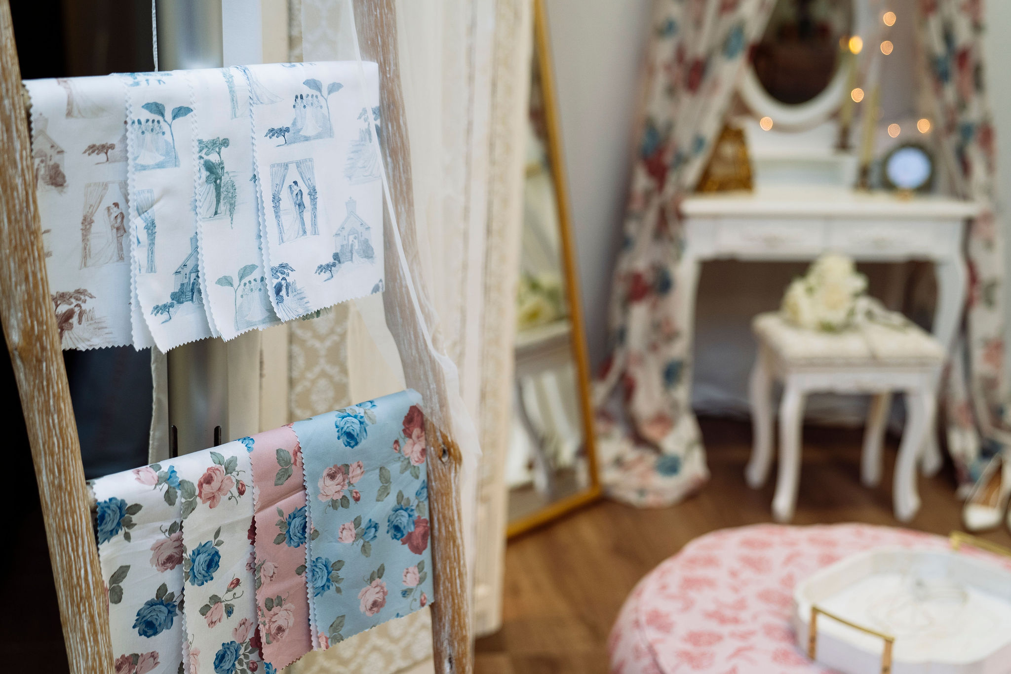 Two rows of patterned fabric swatches hang on a wooden ladder, one with house sketches and one with floral designs. In the background, a floral-decorated vanity mirror and a pink pouf are visible.