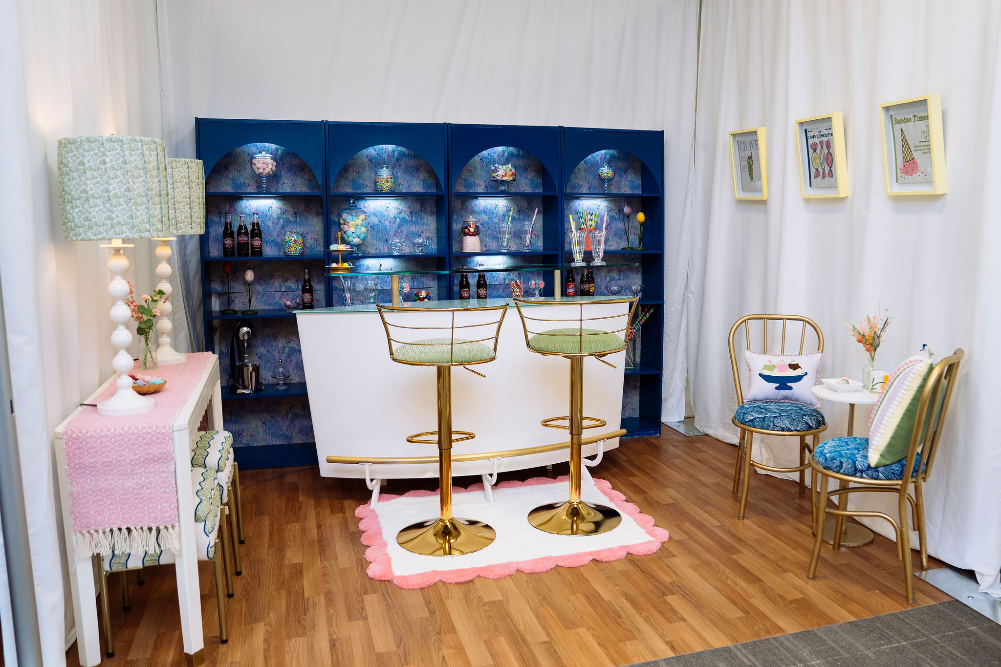 A booth is set up like a candy bar with blue shelves displaying bottles and decor, a white bar with two gold barstools, a small side table with a lamp, and a cafe table with two chairs. Wallpaper, a woven table runner and lampshades are in various blues, pinks and greens.