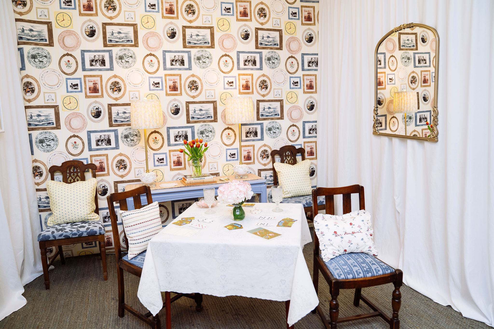 A booth is styled like a dining room with a small table set surrounded by wooden chairs with striped and floral cushions. The wallpaper includes patterned plates and framed photos. A vase of flowers sits on the table, and a mirror hangs on the right wall.