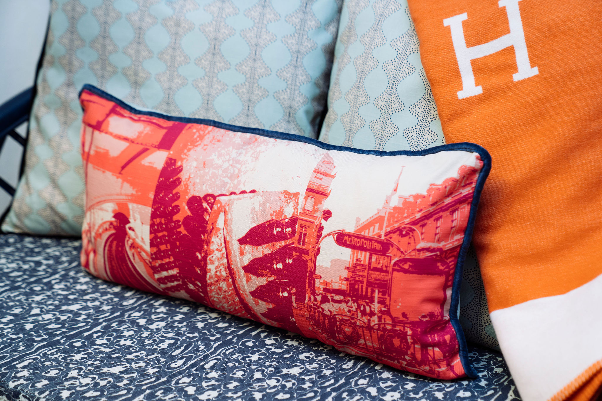 A decorative pillow with a red and orange cityscape design rests on a patterned couch, flanked by a light blue cushion and an orange blanket.