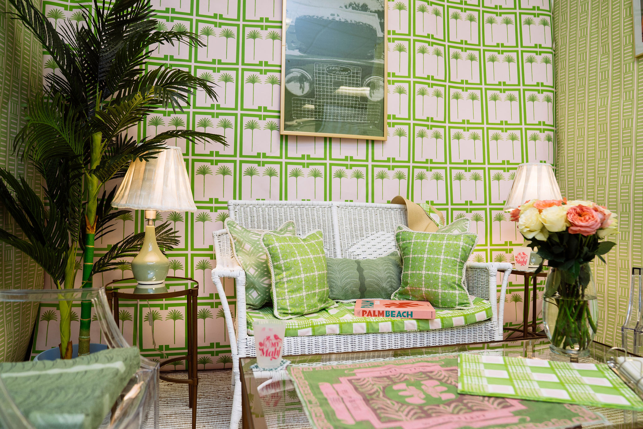 A bright living room with green and white palm-patterned wallpaper, a white wicker sofa with green patterned cushions, a palm plant, two lamps, books, a bouquet of flowers, and a framed car weaving on the wall.
