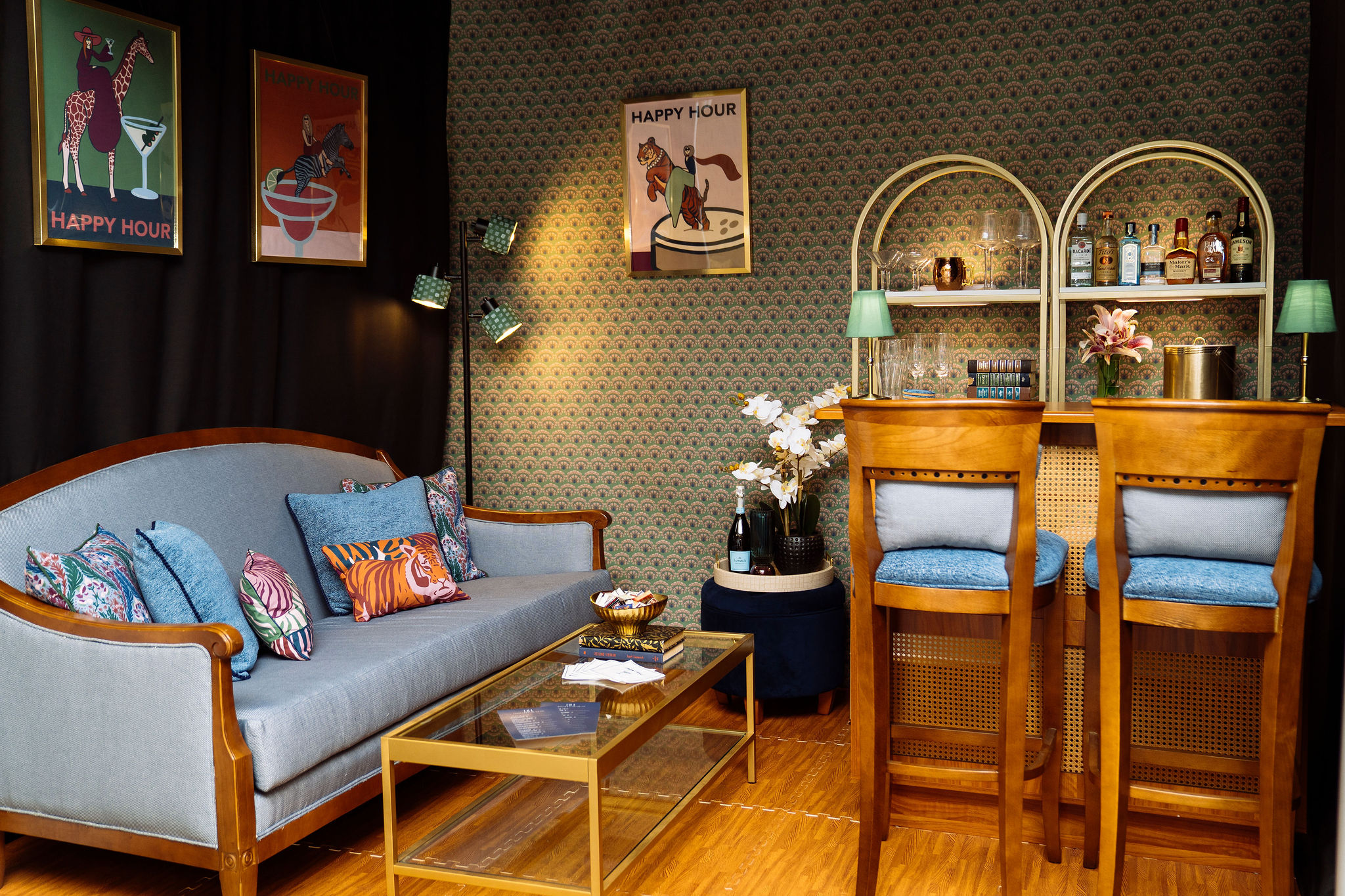 A booth is styled like a bar area with a light blue sofa, colorful pillows, a glass coffee table, and a wooden bar with two tall chairs. The walls have patterned wallpaper and retro “Happy Hour” art, with flowers and lamps as decor.