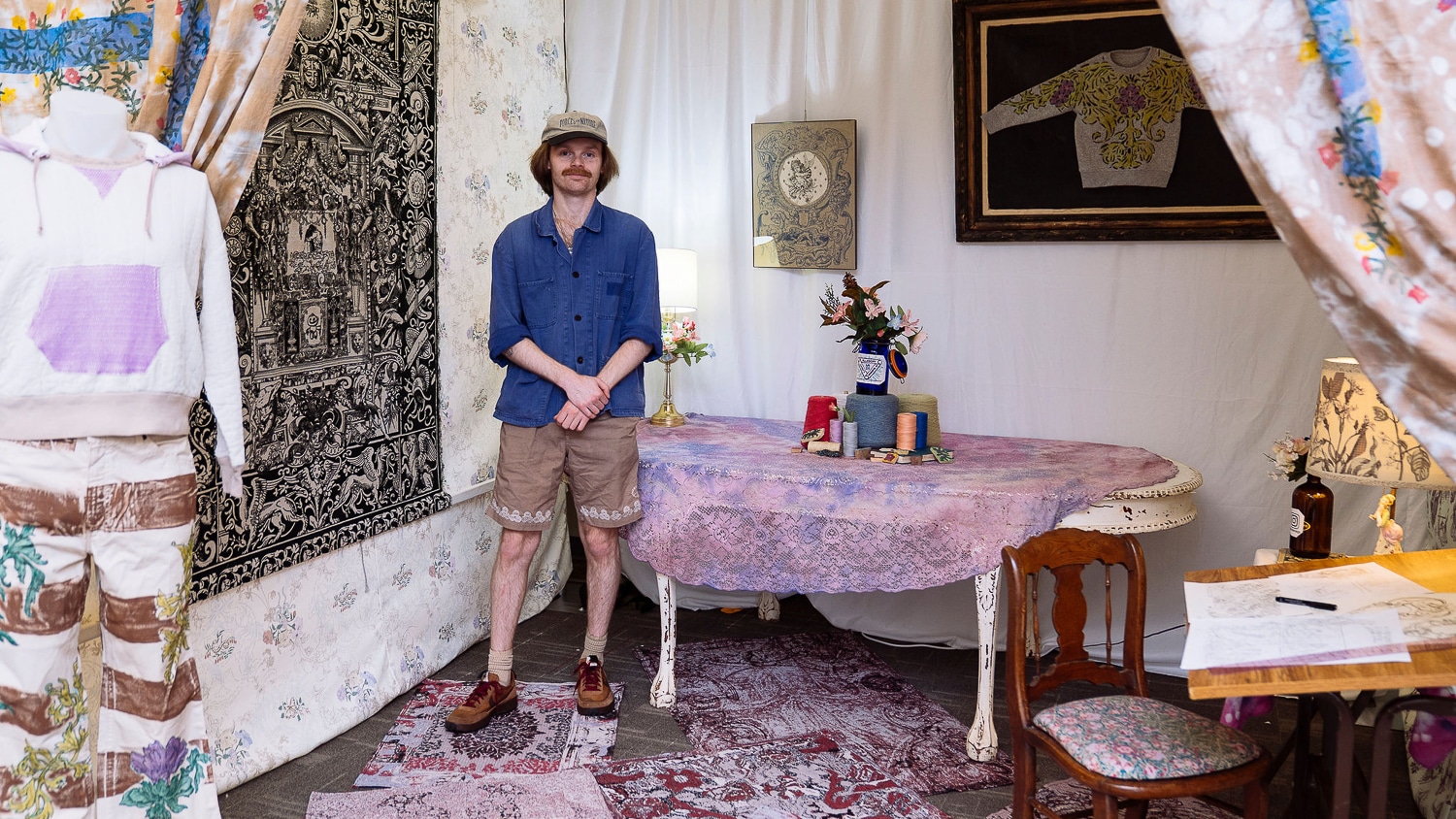 Matthew Riddle stands amid his capstone collection wearing a dark blue button up, khaki shorts and a beige baseball cap. The room is full of eclectically patterned tablecloths, tapestries, rugs and clothing.