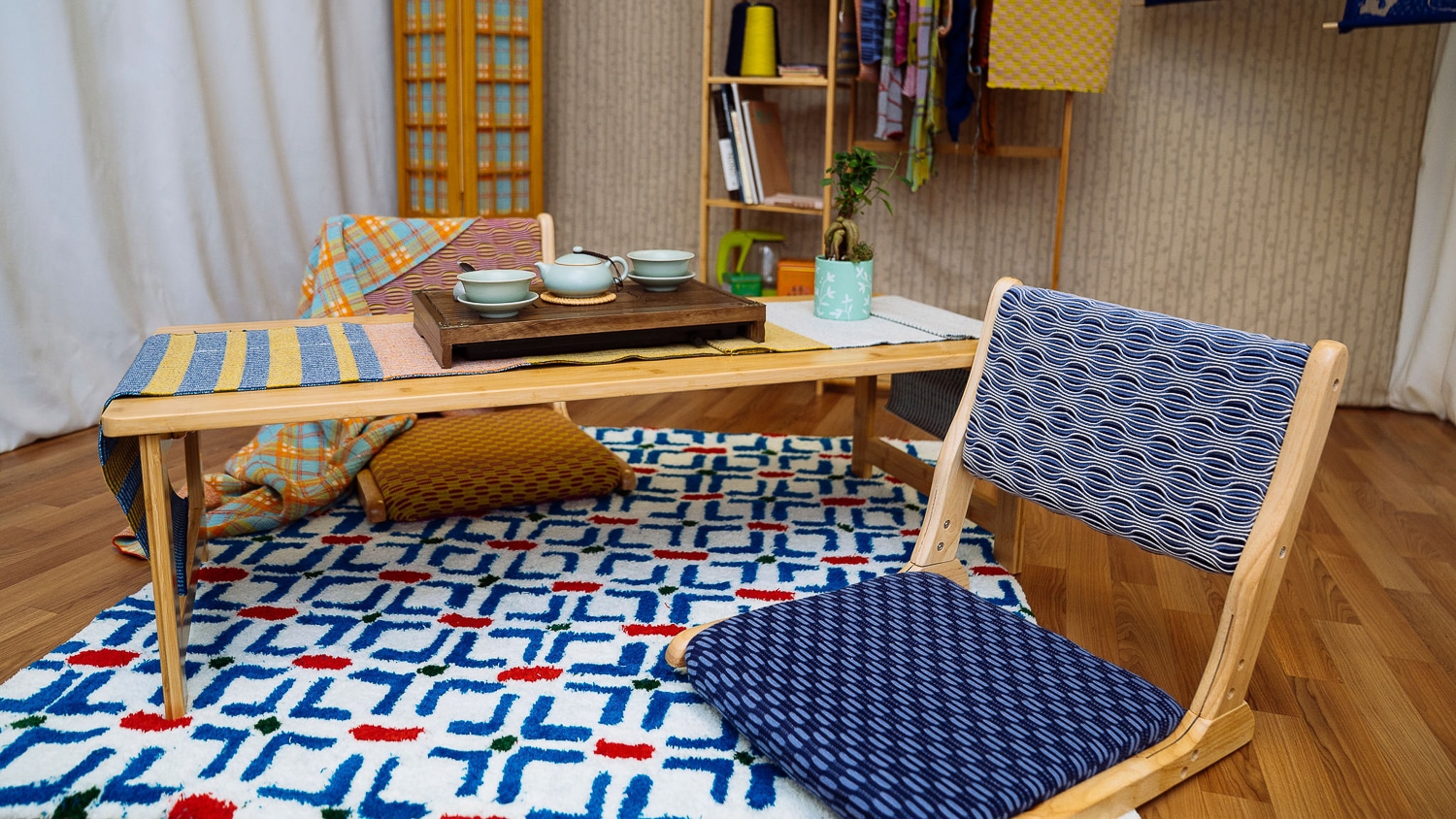 A short table and chairs sit on a white rug with a pattern of blue crosses. One chair has dark blue fabrics, and the other has warmer oranges and a plaid blanket thrown over it. The table is set with a light blue tea set.