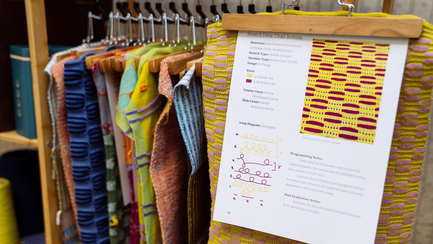 A collection of brightly colored fabrics designed and created by Shayleigh Larsen for her capstone collection hang in a small wardrobe, and a piece of paper displays information for a yellow and red woven design.