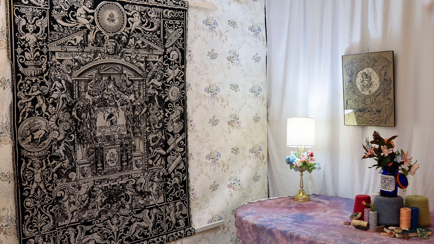 A corner of the room designed by Matthew Riddle. A large tapestry covers one wall, with a dark base and complex scenes woven in white. A small lamp sits on a table with a lacy, hand-dyed tablecloth.