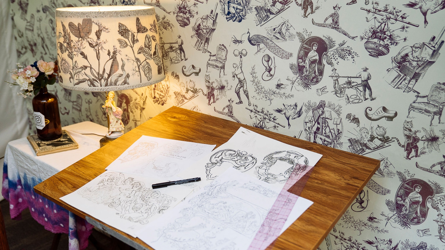 Sketches of detailed, vintage-inspired images are displayed on a wooden table. A lamp shade and wallpaper share the same design: simple black drawings of plants, people and animals evenly dispersed on a white background.