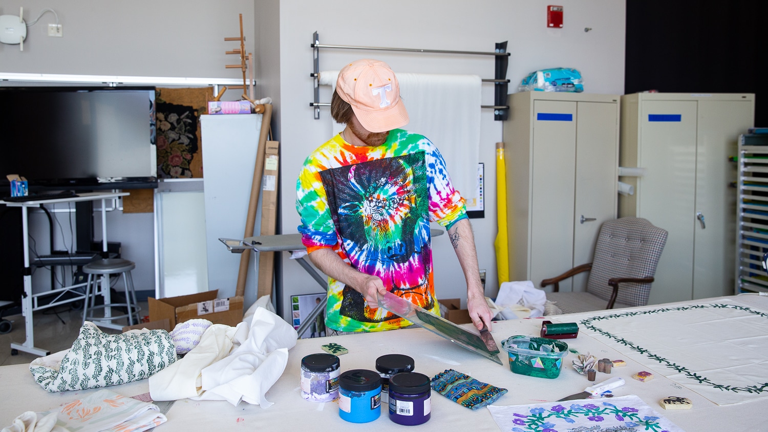 Matthew Riddle wears a tie-dye t-shirt while he works on black prints in the Screen Printing Lab. Jars of ink and various fabrics are splayed on the table in front of him as he makes a swipe of ink.